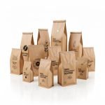 A natural kraft **compostable coffee bag** with a PLA valve and zipper, standing on a coffee shop counter.