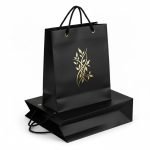 A luxury matte **black paper bag** with a gold foil stamped logo and black rope handles.