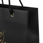 A luxury matte **black paper bag** with a gold foil stamped logo and black rope handles.