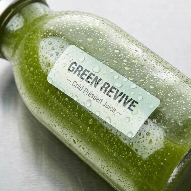 A roll of custom food labels with vibrant graphics being applied to glass juice bottles.