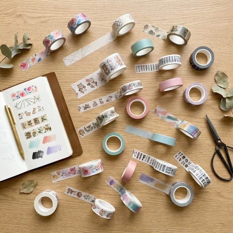 Several rolls of custom washi tape with various colorful floral and geometric patterns stacked on a wooden desk.
