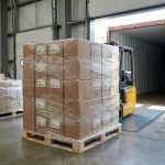 A roll of clear stretch film being wrapped around a pallet of cardboard boxes in a warehouse setting.