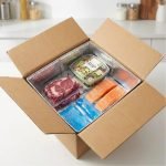 A silver insulated box liner being placed inside a brown corrugated shipping box filled with fresh meat and ice packs.