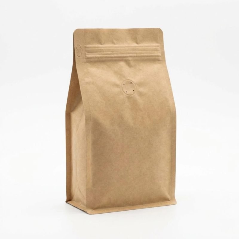 A natural brown compostable coffee bag with a flat bottom and a circular PLA degassing valve standing next to coffee beans.