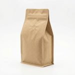 A natural brown compostable coffee bag with a flat bottom and a circular PLA degassing valve standing next to coffee beans.