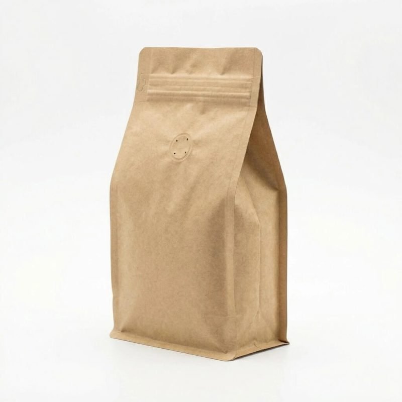 A natural brown compostable coffee bag with a flat bottom and a circular PLA degassing valve standing next to coffee beans.