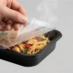 A roll of easy peel lidding film being applied to a plastic food tray containing noodles and meat.