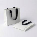 A black luxury tote paperboard gift bag with a gold hot-stamped logo and white grosgrain ribbon handles.
